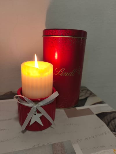 The image shows a close-up of a lit candle in a red holder, with a red Lindt tin in the background. The candle is a thick, white pillar with a yellow flame. It is in a small, fluted red holder that is tied with a white ribbon. A small, white star-shaped ornament hangs from the ribbon. The Lindt tin is red and appears to be cylindrical, with the Lindt logo embossed in gold script on its side. The background is a neutral-colored wall, and the objects are placed on a surface with a patterned tablecloth. The lighting suggests an indoor setting, likely in the evening or a dimly lit room, given the prominence of the candle's glow. The overall mood is warm and festive, possibly associated with holidays or a cozy atmosphere. There is no indication of people, specific weather, or external location cues beyond the mention of Passignano sul Trasimeno, Italy.