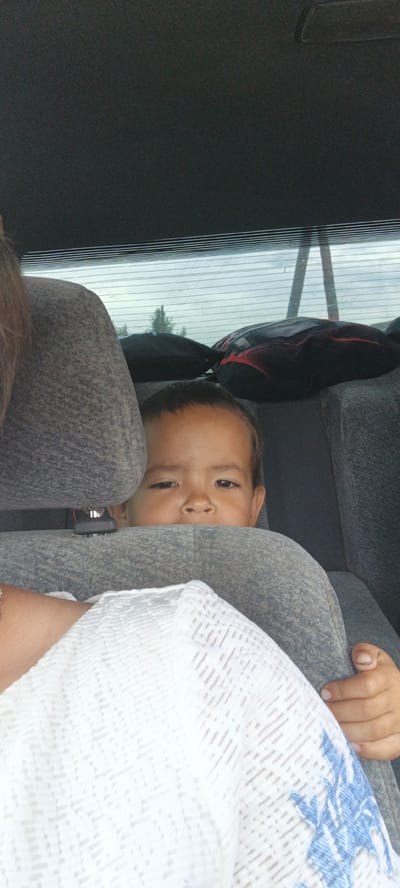 A young child is peering over a car seat in the back of a vehicle. The child has dark hair and brown eyes, and appears to be looking directly at the camera. The child's face is mostly visible, with their eyes and nose prominent. The background shows the interior of the car, including the headrests and the rear window, through which trees and a cloudy sky are visible. A person's arm and hand are visible in the foreground, resting on the car seat, and a white garment with blue patterns is also present. The image appears to have been taken from a low angle, looking up at the child.