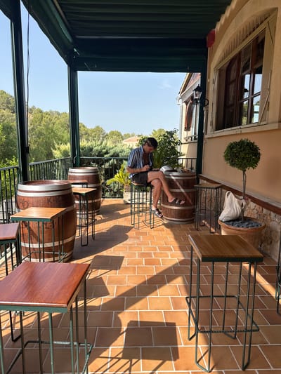 The image depicts an outdoor covered patio, possibly a bar or restaurant area, with a view of lush green hills under a clear blue sky. The patio floor is tiled with reddish-brown square tiles, and there are several wooden wine barrels repurposed as tables. Surrounding these barrel tables are tall, industrial-style bar stools with green metal frames and wooden seats.
In the center of the scene, a man with dark hair, wearing a striped blue and white short-sleeved shirt and dark shorts, sits on a stool. He is looking down at his phone, holding it in his left hand. He is also wearing dark sandals. To his right, a large terracotta pot contains a small, neatly trimmed green tree. A light-colored canvas bag rests on the ground next to the pot.
The patio is covered by a dark green awning, supported by dark green metal posts. A black metal railing runs along the edge of the patio, separating it from the landscape beyond. The building to the right has a cream-colored stucco exterior and features a large wooden-framed window with multiple panes. A black wrought-iron lantern is mounted on the wall near the window. The sunlight casts strong, angular shadows across the tiled floor, indicating a bright, sunny day. The overall ambiance is relaxed and outdoor-oriented, consistent with a Mediterranean setting. The presence of wine barrels suggests a possible winery or establishment with a focus on wine. The city of Gènova, Spain, is known for its coastal location and proximity to vineyards, making such an outdoor seating area plausible.