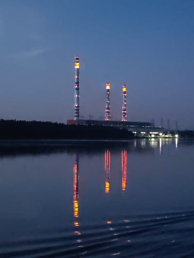 The image shows a power plant at dusk, located on the banks of the Volga River in Konakovo, Russia. Three tall chimneys, illuminated by red and yellow lights, dominate the skyline. The power plant building itself is visible behind the chimneys, with lights also illuminating its facade. The river in the foreground reflects the lights of the chimneys and the building, creating a shimmering effect on the water's surface, which is disturbed by gentle ripples. A dark line of trees separates the power plant from the river. The sky is a deep blue, characteristic of twilight. There are no people or animals visible in the image.