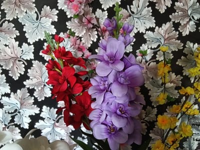 The image shows a close-up view of several artificial flower arrangements in front of a black background with a repeating pattern of stylized grey and white leaves. From left to right, there are branches of red flowers, a large cluster of purple flowers, and smaller clusters of yellow flowers. In the lower left corner, a portion of a white flower is visible. The lighting is even, and the focus is sharp on the flowers. The scene appears to be indoors, likely a decorative arrangement. There are no people or actions depicted. This image is not in Drahichyn, Belarus, as there are no geographical markers or context to suggest this location.