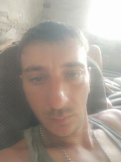 A close-up, low-angle, selfie-style photograph shows a man with fair skin and dark hair, looking downwards. He is wearing a grey tank top and a silver chain necklace. He appears to be indoors, with a textured, light-colored wall visible in the upper left corner and a bright light source emanating from the upper right. The man is positioned on a grey, textured surface, possibly a chair or couch. The background is blurred and out of focus. The specific location within Shpola, Ukraine, cannot be determined from the visual information provided.
