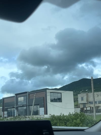 The image shows a view from inside a vehicle, likely a car, capturing an overcast sky filled with gray clouds over what appears to be a residential or commercial area. In the background, a green mountain or large hill is visible. A tall, slender utility pole and power lines run along the roadside. In the middle ground, several modern buildings are seen, featuring boxy structures and large windows. Some of the windows have stacks of materials, possibly construction materials, visible inside. The composition suggests the location might be in or near the city of Gorovani, Georgia, given the presence of modern architecture and the mountainous backdrop.