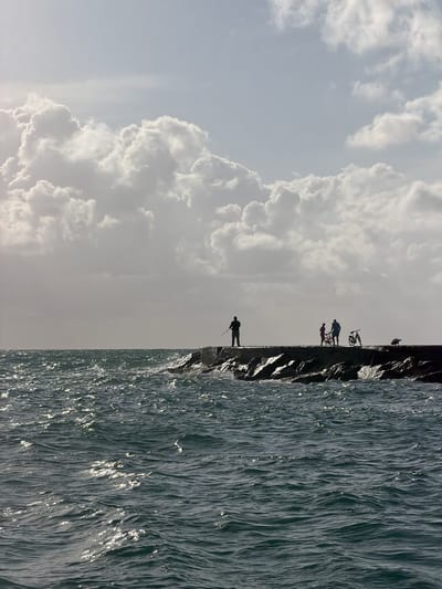 Here is a forensic description of the image:
The image depicts a scene in Bal Harbour Village, Florida. A breakwater structure divides the frame, with the ocean on the right and a cloudy sky on the left. Several individuals are silhouetted against the sky on the breakwater. One person is fishing at the end, while a group with bicycles stands further back. The ocean exhibits wave action and a rippled surface, reflecting light. The sky features prominent cumulus clouds.