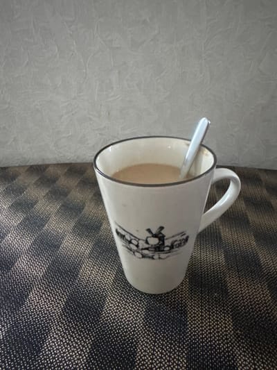 A close-up shot focuses on a white ceramic mug filled with a light brown beverage, likely coffee or tea, with a silver spoon resting inside it. The mug features a black rim and a decorative illustration of a windmill and houses. The mug is positioned on a dark, textured placemat with a woven pattern that reflects light. In the background, a light-colored wall with a rough, textured surface is visible. The image does not provide any information about the city of Usinsk, Russia, nor does it depict any people or actions.