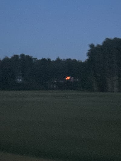 This image depicts a dimly lit scene, likely at dusk or dawn, given the soft, blue sky and the presence of faint, orange lights. The primary subjects are the indistinct silhouettes of trees against the sky and a large, out-of-focus green mass that occupies the right side of the frame.
Nestled within the trees, there are a few small, bright orange specks, which appear to be artificial lights. These lights are clustered together, suggesting they might be from a distant building, a campfire, or perhaps even fireworks. The trees themselves form a dense, dark line across the middle ground, with their jagged edges softened by the low light. The green mass on the right is smooth and featureless, possibly a large structure or a densely vegetated area seen from a distance, though its scale and nature are unclear due to the lack of detail and the blurriness.
The overall impression is one of quiet and possibly remoteness, with the faint lights hinting at human presence or activity in the distance. There is no visible text in the image. The scene appears to be outdoors, given the presence of trees and sky. The atmosphere is calm, with no indication of wind or strong weather.