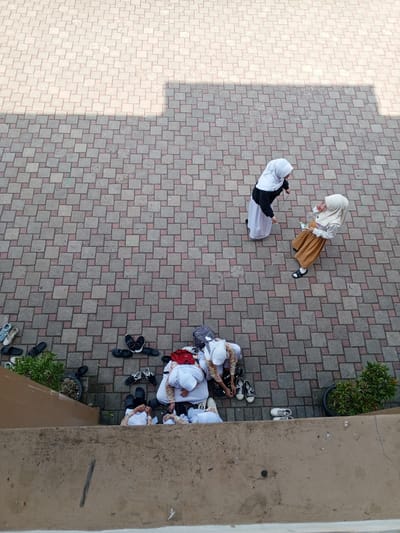An overhead, high-angle shot captures a group of individuals, appearing to be children or young adolescents, gathered on a paved outdoor area. The paving is composed of square tiles arranged in a pattern of light and darker grey shades, with some reddish-brown tiles interspersed. The scene is divided by a diagonal shadow cast from an unseen structure, creating a contrast between a brightly lit area and a shaded section.

In the upper portion of the image, two individuals are standing. One, wearing a white hijab and a black and white garment, interacts with another person wearing a light beige hijab and an orange skirt over white trousers, who is holding a drink and a small white object.

In the lower portion of the image, a larger group of individuals are seated on the ground, many of whom are wearing white hijabs and light-colored clothing. They are in the process of removing or organizing their footwear. A variety of shoes, including sandals, flip-flops, and sneakers, are scattered around them. Some of the individuals are actively engaged in taking off or putting on shoes. A backpack and other bags are also visible near the seated group.

To the left of the seated individuals, a small green plant in a dark pot is visible, alongside several pairs of shoes. To the right, another potted green plant is situated near the edge of the frame. The foreground consists of a concrete ledge or wall, casting a shadow.

The location is described as a paved area in the city of Chutula, Mozambique. The overall impression is one of a group of students preparing to enter or exit a building, possibly a school or place of worship, where removing shoes is customary.