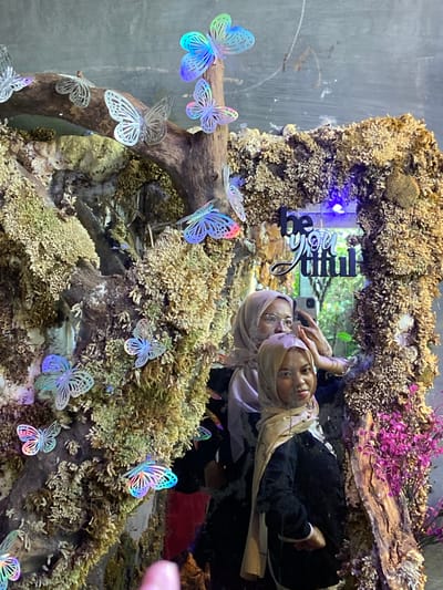 The image depicts two women, appearing to be of Southeast Asian descent, posing for a selfie in front of a decorative display. The display features a rustic, moss-covered structure with numerous iridescent, butterfly-shaped ornaments. A portion of a mirror is visible, reflecting the women and the surroundings, which include greenery. Above the women, the words "be you tiful" are mounted on the display in black lettering. The overall setting suggests an indoor environment, possibly a retail space or a themed attraction, as indicated by the artificial nature of the butterflies and moss. There is no explicit indication of the city of Rengat, Indonesia within the visual information provided.