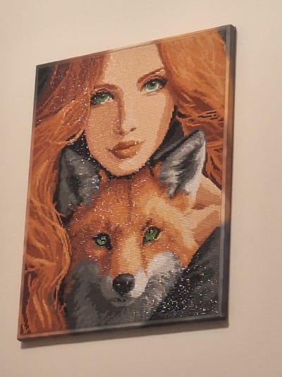 A framed artwork featuring a stylized portrait of a woman with long, wavy, orange hair and green eyes, partially embracing a fox. The artwork has a pixelated or mosaic-like appearance, with individual colored pieces forming the image. Sparkly elements are interspersed throughout the artwork, adding a shimmering effect. The woman's face is in the upper half of the frame, with the fox's face in the lower half, nestled against her. The background is dark. The artwork is mounted on a wall. The location in Mińsk Mazowiecki, Poland, is not determinable from the image.