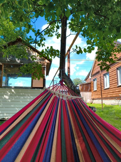 The image depicts a colorful striped hammock suspended between a tree and an unseen support structure, in the yard of a wooden house. The hammock is made of fabric in alternating stripes of red, dark blue, green, and beige. The tree has a thick, rough trunk and is partially obscured by lush green leaves, with branches reaching towards a bright blue sky with scattered white clouds.
In the background, a two-story wooden house with a brown exterior is visible. It has several white-framed windows and a slanted roof. To the left of the house, another smaller wooden structure with a large window reflecting the sky is present. The ground is covered in green grass, with some patches of bare earth and a low wooden fence. The scene appears to be in a rural or suburban setting, possibly a dacha or a country house. The location is identified as Munayka, Russia. There are no people visible in the image.