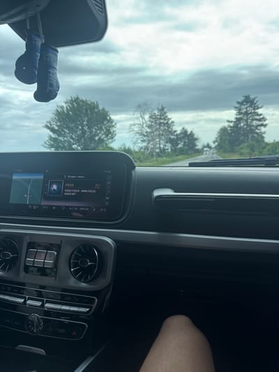 The image shows the interior of a car, likely a Mercedes-Benz G-Wagon, as indicated by the dashboard and the ventilation vents. The infotainment system displays the radio station "Focus" at 99.7 FM, which is likely a Bulgarian radio station. The image is taken during daytime with an overcast sky visible through the windshield. There are trees outside, suggesting a rural or suburban environment. A pair of boxing glove-shaped air fresheners are hanging from the rearview mirror. The climate control is set to 22 degrees Celsius, with A/C enabled. The location is suggested as Bazan, Bulgaria.