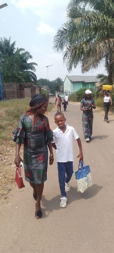 The image captures a vibrant, sunlit scene in Ohafia, Nigeria, showing several people walking along a road. In the foreground, a woman with dark skin, wearing a patterned, knee-length dress with ruffled sleeves, a black head covering, and open-toed sandals, smiles while carrying a red purse and a phone. Beside her, a young boy with dark skin, dressed in a white polo shirt, blue patterned pants, and white sneakers, also smiles, holding a blue and white geometric patterned tote bag with "TLY" faintly visible on its tag.
Further down the road, another woman in a patterned jumpsuit and white cap walks, while behind her, a woman carries a yellow jerrycan on her head. In the background, two men and a small child are walking away from the camera. The road appears to be dirt or unpaved, bordered by abundant green foliage, including several tall palm trees. A light green building with a dark roof is visible in the distance, alongside other structures and street lights. The clear sky and strong shadows indicate a bright, sunny day. The expressions of the woman and boy in the foreground convey a cheerful mood.