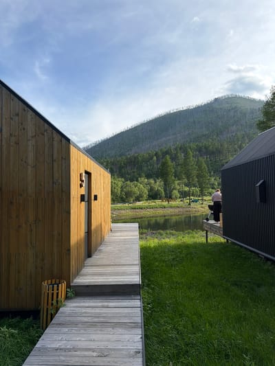 The image depicts a serene outdoor scene in Bol'shoe Goloustnoe, Russia, likely at a lakeside resort. In the foreground, two modern cabins are visible, constructed from wooden planks and corrugated metal. A wooden walkway connects the two structures and leads towards the water. The cabin on the left is made of light-colored wood, with a dark-framed door. Next to it, a woven cylindrical trash bin stands against the wooden wall. The cabin on the right is clad in dark corrugated metal. A person, wearing a light pink top and dark pants, stands on the deck of this cabin, facing away from the camera, tending to a charcoal grill.
The middle ground features a calm body of water, reflecting the sky and the lush green vegetation along its banks. The water is bordered by a grassy area and a dense forest. In the background, a steep, tree-covered mountain rises, with some areas showing evidence of past fires, indicated by a sparse scattering of dead trees on the upper slopes. The sky is partly cloudy with soft, diffused sunlight, suggesting it is either early morning or late afternoon. The overall atmosphere is tranquil and natural.