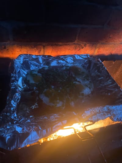 A close-up, overhead shot shows food being cooked in aluminum foil over a fire. The food appears to be some kind of baked dish, possibly potatoes or a casserole, with visible white and green toppings, and possibly some red pieces. The aluminum foil is crimped around the edges to contain the food. The fire below is bright orange and yellow, casting a warm glow on the foil and the brick structure behind it. The brick structure is rough and appears to be part of a fireplace or outdoor oven. The background is dark, with the light of the fire being the primary illumination. The image does not provide enough information to determine the specific location in the city of Chulym, Russia.