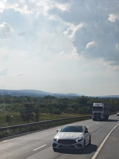 In Novosemeykino, Russia, under a bright, cloudy sky, a white Mercedes-Benz car with the license plate X410CE763 is traveling on a multi-lane highway. A white semi-truck is driving in the lane to the right of the car. The highway is bordered by a metal guardrail and dense green vegetation on the left. In the distance, rolling hills covered in trees and scattered buildings form the horizon. The scene depicts a typical highway commute in a rural or semi-rural area.