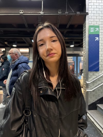 A woman with dark hair, wearing a black leather jacket and white earphones, smiles at the camera. She stands on a subway platform, with a blue and green directional sign above. An elderly man in a blue jacket and face mask stands nearby, along with other people in the background. A subway train is visible behind the woman. The platform features tiled walls and metallic railings.