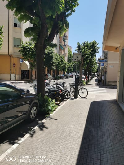 This is a street scene in Follonica, Italy, on a sunny day. The foreground is dominated by a dark-colored car parked along the curb. Adjacent to the car is a sidewalk paved with square tiles. Several motorcycles and a bicycle are parked on the sidewalk, some leaning against a tree trunk. Beyond the parked vehicles, a street stretches into the distance, lined with buildings. A pedestrian crossing is visible further down the street. Signs indicate parking regulations and a ticket vending machine. Some people are visible walking on the sidewalk in the middle ground. Trees with green foliage line the street, casting shadows on the sidewalk and road. The sky is clear and blue.