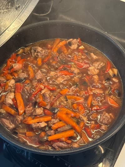 An overhead, close-up view captures a hearty stew simmering in a dark, round pan on a black induction or ceramic stovetop in a kitchen in Shirkovo, Russia. The pan is filled with a rich, dark brown liquid, gently bubbling, suggesting it is actively cooking. Faint steam is visible rising from the dish.

The stew contains numerous ingredients: prominent chunks of tender meat, likely beef or pork; vibrant orange carrot sticks cut into elongated pieces; and bright red bell pepper strips. Darker, softer vegetable pieces, possibly eggplant or zucchini, are also mixed throughout the bubbling liquid.

To the bottom left, a clear glass pot lid, covered with condensation, is partially visible, resting next to the pan. On the right side of the stovetop, a control panel with white markings is visible. These markings include numbers "0", "1", "3", "5", "8", "10", and "14 P", which likely denote heat levels or timer settings. A circular icon featuring a plus and minus sign, and another small circular icon, are also present on the control panel. The scene conveys the process of preparing a home-cooked meal.