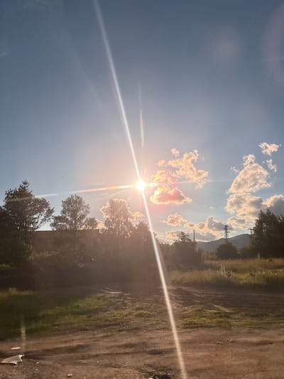 The image depicts a rural landscape under a bright, sunlit sky. The sun is prominently positioned in the upper center of the frame, emitting strong lens flares that streak across the image. Scattered clouds, some illuminated with warm, orange hues, dot the sky. In the foreground, a dirt path winds through grassy terrain. Several trees are visible, silhouetted against the bright sky and bathed in sunlight. In the mid-ground and background, there are indications of human infrastructure: power lines and poles are present, as well as what appears to be a building or structure partially obscured by trees. Distant hills or mountains can be seen on the horizon. The overall impression is of an outdoor scene, possibly in the late afternoon or early morning due to the angle of the sun and the warm lighting. The location is identified as Pirdop, Bulgaria.