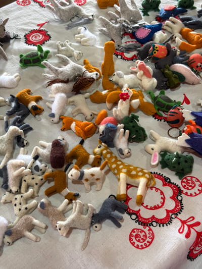 A close-up overhead shot shows a collection of felted animal figurines arranged on a white tablecloth with red embroidered floral patterns. The figurines, predominantly in muted earth tones and grays, depict various animals such as dogs, cats, rabbits, frogs, a turtle, a fox, a bird, a giraffe, and a kangaroo. Some of the figurines are attached to keychains. The arrangement appears to be a display or a collection of handmade crafts. The location is identified as a market stall in the city of Кызыл-Туу, Kyrgyzstan.