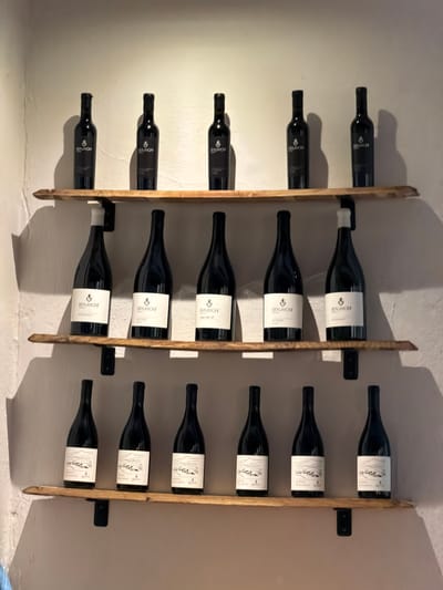 The image displays a wall-mounted wine rack holding 18 dark-glass wine bottles, arranged horizontally in three vertical columns against a light-colored wall. The rack is constructed from vertical wooden slats, which secure the bottles, and is attached to the wall by black metal brackets.
The bottles feature two distinct label designs. The bottles in the leftmost and rightmost columns have predominantly black labels with white text. The word "BEAUMONT" is clearly visible at the top of these labels, often accompanied by "ESTATE" and "SOUTH AFRICA." Other visible text fragments include "B.O.R" and "BOSCHETAL," and some labels may show vintage years like "2021."
The bottles in the middle column feature white labels with black text and a prominent illustration depicting a landscape of mountains or hills. "BEAUMONT" is also present at the top of these labels. Additional text, such as "SAUVIGNON BLANC" and specific vintage years (e.g., "2022"), can be faintly seen on some of these white labels.
The lighting appears to be artificial, casting distinct shadows of the bottles and the rack onto the wall, indicating an indoor setting. The organized display suggests a dedicated space for wine storage, such as a cellar or tasting room. Given the context of Theewaterskloof Nu, South Africa, the "Beaumont" wines are likely from a winery in that region.