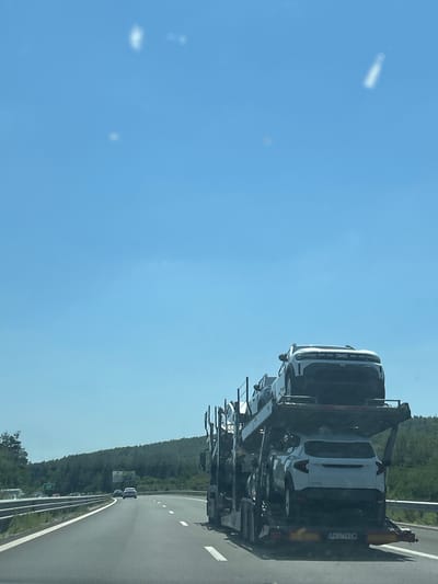 An eye-level, medium shot captures a car carrier truck transporting new vehicles on a highway in Rajana, Bulgaria. The truck, with two levels of cars, is positioned on the right side of the frame, obscuring a significant portion of the view. The top level of the carrier holds a white SUV with a distinctive front grille and headlights. Below it, another white SUV is visible, its rear facing the camera. The highway stretches ahead, curving to the left, with other vehicles and a green hillside in the distance. The sky is a clear, bright blue with a few blurry white spots, possibly lens flares or clouds. The overall scene depicts a mundane transportation event on a sunny day.