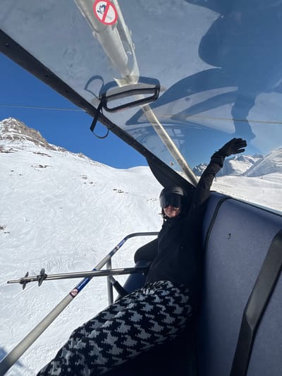 A single individual is observed inside a covered chairlift cabin, ascending a snow-covered mountain slope in Klösterle, Austria. The person is seated, wearing a black ski helmet, dark ski goggles, a black ski jacket, black gloves, and black and white houndstooth pattern ski pants. Their left arm is raised above the head, hand open, and a smile is visible on the lower portion of their face, directed towards the camera. The chairlift cabin features transparent plastic roofing, dark blue upholstered seating, and a metallic safety bar with an attached silver ski pole. A red and white circular prohibitory sign, depicting a skier with a diagonal line through it, is affixed to the interior roof frame. The surrounding environment consists of expansive snow-covered mountain peaks with visible ski trails and a clear blue sky.