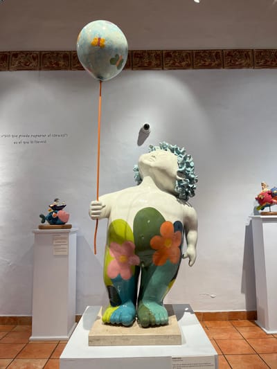A whimsical ceramic sculpture of a figure with floral designs on its body holds a balloon on a stick. The figure has a head of blue curls and a contemplative expression. A small fish-like sculpture sits on a pedestal below. The setting appears to be an art gallery, with text on the wall beside the balloon and tiles running along the wall's edge. A security camera is visible on the wall above the sculpture.