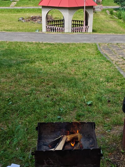 The image captures a scene in Sosny, Ukraine, with a focus on a barbecue grill in the foreground. The grill, a dark metal brazier, contains burning wood, with flames licking at the logs. The surrounding area is covered in green grass, suggesting an outdoor setting.
In the background, a small, open-sided structure with a red roof stands prominently. It's built of light-colored bricks and features arched openings with decorative wooden railings. Beyond the structure, more grassy slopes and a paved pathway are visible.
The overall impression is one of a casual outdoor gathering or a preparation for a barbecue. The natural lighting suggests it is daytime, likely with mild weather. The setting appears to be a park or recreational area. There is no visible text in the image.