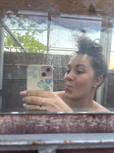 The media file shows a mirror reflection of a young woman in what appears to be an outdoor shower. She has dark, curly hair pulled up with a gold heart-shaped clip. Her face is angled downwards, and she is holding a smartphone with a floral-patterned case, suggesting she is taking a selfie. She is wearing a silver chain necklace. On her finger, she wears two rings, one of which appears to be a simple band, and the other a thin, textured band. Her fingernails are painted a pale pink.

The background in the reflection shows a showerhead with water spraying, and beyond that, greenery with leaves, possibly from a vine or tree, suggesting an outdoor setting. There are also what appear to be industrial metal structures reflected in the mirror, possibly part of a shower enclosure or surrounding infrastructure. The lighting suggests it is daytime. The overall mood seems relaxed and casual. There is no visible text in the image. The location context is Vaklino, Bulgaria.