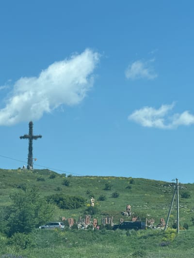 This image captures a panoramic view of the Saghmosavan Alphabet Monument and the Cross of the Fallen in Saghmosavan, Armenia. In the foreground, a grassy hill slopes upwards, dotted with green bushes and scattered yellow wildflowers. Several cars, including a silver minivan and a black SUV, are parked amidst the vegetation.
Further up the hill, large stone letters, forming Armenian script, are visible. These letters appear to be part of a monument, with some displaying weathered surfaces and graffiti. In the background, a towering metallic cross, known as the Cross of the Fallen, stands prominently against a clear blue sky with scattered white clouds. The cross is constructed from dark metal and has a rustic, unfinished appearance. A small flag, possibly red and white, is affixed to its base.
The environment suggests a sunny day with a gentle breeze, as evidenced by the white, puffy clouds. The overall scene conveys a sense of history and cultural significance, with the monuments serving as landmarks against the natural backdrop of the Armenian landscape.