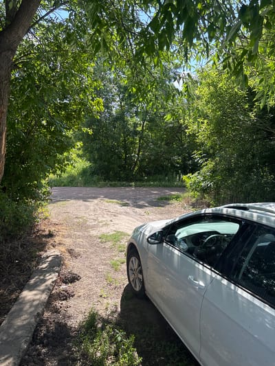 A white car is parked on a dirt road surrounded by trees and greenery. The car is a hatchback, with its sunroof open. The road appears to be unpaved and leads into a wooded area. The sky is blue with some clouds. The image was taken on a sunny day. The location is Pereiaslavske, Ukraine.