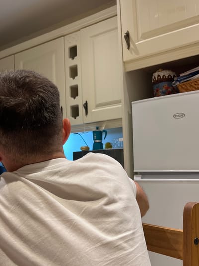 A man, seen from behind, is seated at a wooden table in a kitchen in Balbriggan, Ireland. He is wearing a white t-shirt. The kitchen has cream-colored cabinets and a white refrigerator with a "THOR" logo. On a shelf above the refrigerator, a decorative cake and a woven basket are visible. A turquoise coffee maker sits on a counter in the background. The scene is indoors and appears to be domestic.