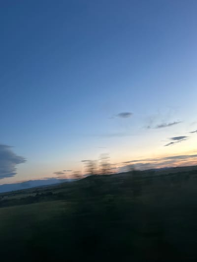 A landscape scene captured in Smilets, Bulgaria, reveals a sky transitioning through sunset. The upper left is a clear blue, slowly merging into bands of yellow, orange, and grey-toned clouds as the sun approaches the horizon. Below this, a blurred line indicates the top of a dark mass, possibly representing land or water reflecting the diminishing light. The ground vegetation or landscape is nearly impossible to ascertain. There are no people visible in the image. The location is Smilets, Bulgaria.