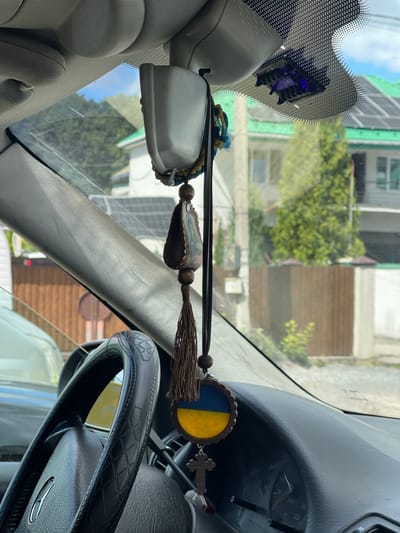 The image is a close-up, interior view of a car in Konopnytsia, Ukraine, focusing on the rearview mirror area and the dashboard. A black steering wheel with a patterned grip is visible on the left. Hanging from the rearview mirror assembly are several car charms. These include a decorative beaded tassel, a small oval pendant depicting an image of a religious figure, and a circular pendant with the blue and yellow colors of the Ukrainian flag. Below the Ukrainian flag pendant, a small wooden Christian cross is also hanging. The car's dashboard and windshield are visible, with a view of houses and trees through the windshield. The lighting suggests it is daytime.