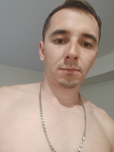 A close-up, head-and-shoulders selfie shows a shirtless man with short brown hair and light stubble. He is wearing a silver chain necklace. The background is a plain, light gray wall. The image was taken in Shkurupiivka, Ukraine.
