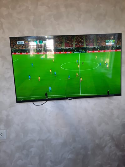 The image shows a flat-screen television displaying a soccer match. The score displayed on the screen is "47:27 COL 0 0 COD", indicating 47 minutes and 27 seconds into the game with the score tied at 0-0 between "COL" and "COD". The television is mounted on a wall with a textured, greyish finish. A black cable is visible hanging from the bottom of the television. The environment appears to be an indoor setting, likely a living room. The television is positioned in the center of the frame, dominating the view. There are no discernible elements in the image that would allow for the specific identification of the location as Burma-Suu, Kyrgyzstan. The scene is static and depicts a typical viewing experience of a live sports broadcast.