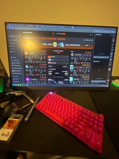 This is a close-up, eye-level shot of a gaming setup. The primary subject is a computer monitor displaying a game interface, likely a first-person shooter, with player statistics and match information. A red mechanical keyboard with RGB lighting is prominently placed in the foreground, partially obscuring a pack of Winston cigarettes and a pen. The scene appears to be indoors, with a dark, reflective surface covering the desk. The monitor shows a match between two teams, "team_Nies" and "team_SPRAYJUTSU", with player stats like win rate, kills, K/D ratio, and ADR displayed. There is no indication of the city of Pobiedziska, Poland, within the image content.