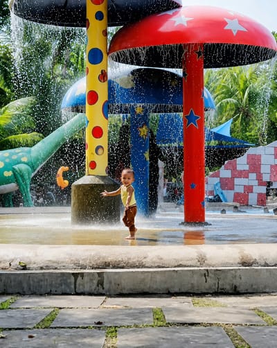 A young child, possibly a toddler, is joyfully playing in a shallow water area of an outdoor water park. The child is barefoot and wearing a yellow sleeveless shirt and brown pants. They are smiling and reaching out with their right hand. The water is splashing around them from overhead mushroom-shaped structures. One mushroom structure is yellow with red and blue polka dots, another is blue with yellow stars, and a third is red with white stars. A green dinosaur-shaped slide is visible in the background to the left. The water park appears to be surrounded by lush green palm trees and other vegetation, suggesting a tropical or sub-tropical location. The ground in the foreground consists of concrete slabs with grass growing between them. The overall impression is one of a fun and lively atmosphere for children. This scene is located in the Pantai Cermin Kiri area of Indonesia.