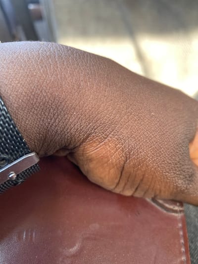 In Zankan, Nigeria, the scene presents a close-up, downward-facing view of a dark-skinned human left hand and wrist. A watch with a black mesh band and a silver-colored clasp is present on the wrist. The hand is resting, with its palm facing down, on a reddish-brown object that appears to be a leather-bound item, possibly a book, notebook, or wallet, showing some subtle texture and faint scuffs. The background beyond the hand and object is out of focus, characterized by diffuse light and shadow.