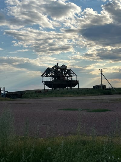 The image depicts an abandoned industrial structure, possibly a former processing plant or factory, set against a dramatic sky filled with dramatic clouds illuminated by rays of sunlight. The structure is skeletal and in disrepair, made of metal and wood, with a distinctive multi-tiered design. In the foreground, there's a flat, gravelly area. To the left, a blue tractor trailer is parked. The background features a grassy field with a small, dilapidated shed and a utility pole. The overall impression is one of rural decay and desolation. The specific location in Bayagiz, Kazakhstan, is not visually identifiable from the image itself, but it is presented as the setting.