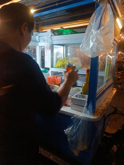 A person is seen from behind and to the left, wearing a dark shirt, standing at a food stall in Luwoo, Indonesia. The food stall appears to be a mobile cart with a glass display case. Inside the display, there are yellow noodles and other food items. An orange container and a bottle with liquid are visible on the counter. A plastic bag hangs from the top of the stall. The scene is lit by artificial lights, suggesting it is evening or nighttime. A motorcycle is parked to the right of the stall.