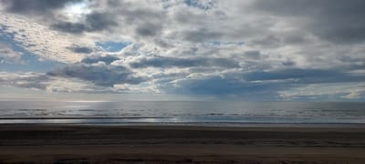 This image depicts a serene beach scene with the ocean meeting the shore under a cloudy sky. The foreground is dominated by dark sand, with subtle tire tracks indicating recent activity. A thin strip of water lies between the sand and the main body of the ocean, reflecting the muted light. The ocean itself is calm, with gentle ripples stretching towards the horizon. The sky is filled with a dramatic display of clouds, ranging from bright white formations to darker, more ominous gray ones. Patches of blue sky peek through the cloud cover, and the sun is partially visible, casting a soft glow on the water. The overall impression is one of a peaceful, somewhat overcast day at the coast. The location is the beach of Aleksandrovsk-Sakhalinskiy, Russia.