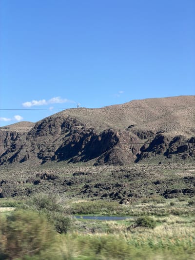 The image shows a vast, arid landscape under a clear blue sky. A rugged, rocky mountain dominates the middle ground, its slopes covered in sparse vegetation and dotted with dark, weathered rocks. Several utility poles and wires are visible traversing the mountainside, suggesting human infrastructure in this remote area. At the summit of the mountain, a small structure with antennas can be seen. In the foreground, a blurred stretch of green vegetation and a small body of water suggest a slightly more fertile area. This scene is likely located near Kok Moynok Eki, Kyrgyzstan, a region known for its mountainous terrain and natural beauty.