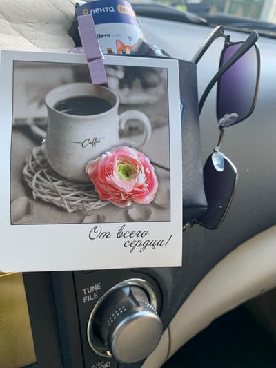 The image shows a close-up view of a car's dashboard in Yurga, Russia. A photograph of a coffee cup with the word "Coffee" written on it, alongside a pink and green rose, is pinned to a dark blue holder with a light purple clothespin. The photograph has the Russian text "От всего сердца!" (From the bottom of my heart!) written below it. A pair of sunglasses with purple lenses is hanging from the holder. To the right of the photograph, a portion of the car's steering wheel is visible. Below the photograph, on the car's stereo system, the text "TUNE FILE" is visible, along with a silver knob. The environment appears to be inside a vehicle, with the dashboard being the primary focus.