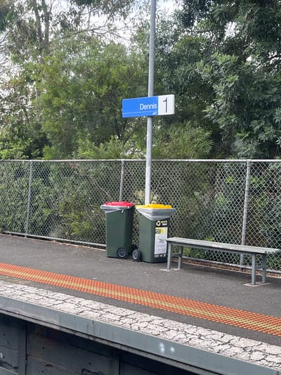 The image displays a train station platform, likely Dennis Station in Northcote, Australia, during daylight hours on an overcast day. On the left side of the frame, a blue and white sign, mounted on a metal pole, clearly indicates "Dennis" with "1" above it, signifying Platform 1. A small Public Transport Victoria (PTV) logo is also visible on the sign.

Adjacent to the sign and pole, two green wheelie bins are placed near a metal bench. The bin closer to the bench has a yellow lid, prominently labeled "Recycle." Below this, a sticker specifies accepted items: "Recycle Paper & Card, Hard Plastic Containers, Steel & Aluminium," and includes a "No Plastic Bags" symbol. The second bin has a red lid, typically used for general waste, although its specific text is not legible.

Behind the station infrastructure, a chain-link fence separates the platform area from a dense background of green trees and foliage. The platform surface is asphalt, featuring a tactile safety strip of orange-yellow raised bumps running parallel to a white line along the edge, indicating the safe distance from the railway tracks. No people are present, and no activity is occurring, depicting a quiet scene of station amenities.