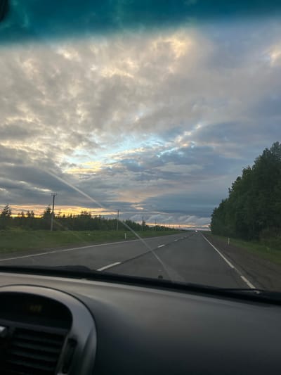 The image depicts a view from inside a car, looking out through the windshield. The car appears to be traveling on a road adjacent to a forested area. The sky is filled with clouds, some of which are illuminated by the setting sun. Telephone poles line the edge of the road. The location is Gorshki, Russia.