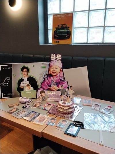 A low-angle, medium shot captures a table laden with merchandise and a cake, likely for a birthday celebration. The background reveals a dark, textured wall meeting a ceiling with square, illuminated glass panels and a pendant light. A dark, cushioned bench is visible behind the table.
Dominating the table are several items featuring a young K-pop artist or idol, seemingly named "Jeon Wonwoo". To the left, a large poster depicts the artist in glasses, holding a cake, with "Happy Birthday" text. Adjacent to it, a smaller cutout shows the artist in a white shirt. A cutout of a baby dressed in a traditional Korean outfit, also featuring the name "Wonwoo", stands prominently in the center.
A tiered cake, decorated with purple and white frosting and topped with small cutouts of the artist and a cartoon cat, sits on the right side of the table. Surrounding the cake are various K-pop merchandise items, including photo cards, keychains, and small plush toys, all featuring the artist. A clear plastic fan is also visible. The overall scene suggests a fan event or personal celebration. The environment does not specifically indicate a location in Serang, Indonesia.
