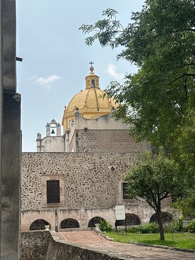 The image captures a historical architectural complex, likely a church or monastery, set amidst lush natural surroundings under a clear daytime sky. The central feature is a building with a prominent, golden-yellow dome topped with a cross, visible against a light blue sky with a few faint clouds. Below the main dome, a smaller bell tower is visible, housing a dark bell.
The building's walls display varied construction materials. The lower right section is composed of rough-hewn, light-colored stones, giving it a rustic, textured appearance. This section includes several arched alcoves or openings and a small, gridded window. Adjacent to this, a wall section made of neatly laid reddish-brown bricks suggests either a different construction phase or architectural style.
Dense green foliage from large trees frames the upper and left parts of the image, indicating a well-treed environment. In the foreground and to the right, a paved path or low stone wall borders a patch of green grass with further shrubs and a small tree. A dark, rectangular sign with white, mostly illegible text, likely an informational plaque, stands near the base of the stone wall on the right.
No people are present, lending a serene and tranquil atmosphere to the scene. The bright, even lighting suggests it is daytime, likely morning or early afternoon. The architectural style, with its dome and bell tower, is characteristic of colonial-era religious buildings found in Mexico, fitting the context of Tepotzotlán.