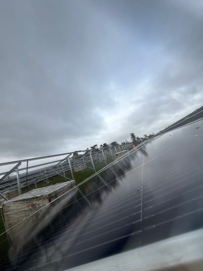 The scene depicts an active solar farm construction site at Baldurgan Hill, Ireland. The perspective is slightly angled, looking across a green, gently sloping field under an overcast, grey sky with a brighter segment visible in the upper right. In the immediate foreground on the right, several dark, rectangular solar panels are partially installed on a metallic racking system, displaying visible moisture droplets on their reflective surfaces. The mid-ground features numerous parallel rows of unpaneled metallic support structures for solar panels, extending into the distance across the grassy terrain. To the left of the installed panels, multiple large, palletized stacks of construction materials, wrapped in clear plastic, rest on the ground. A small, orange, conical object is discernible among the distant racking. A dense tree line forms the background horizon. No individuals are observed within the frame.